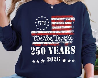 May include: Navy blue sweatshirt featuring a distressed American flag design with the text "1776" above the flag and "We the People" below. The design also includes "250 YEARS 2026" and stars.