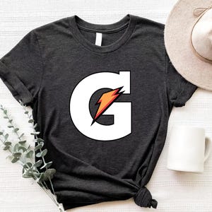 May include: Dark gray t-shirt featuring a large white "G" with a red and orange lightning bolt design. The shirt has a crew neck and short sleeves. A beige hat and white mug are also visible.