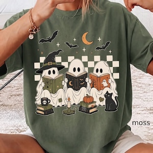 May include: A moss green t-shirt featuring a graphic of three ghosts reading books. One ghost wears a witch hat. The design includes a crescent moon, bats, a black cat, a pumpkin, stacks of books, and a checkered pattern background in cream.