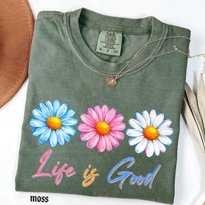 Pode incluir: Camiseta verde musgo com tr&ecirc;s desenhos de flores de margarida em azul, rosa e branco, cada uma com um centro amarelo. A frase "Life is Good" est&aacute; impressa abaixo das flores em uma escrita colorida. Um colar de prata com um pequeno pingente repousa na camisa.