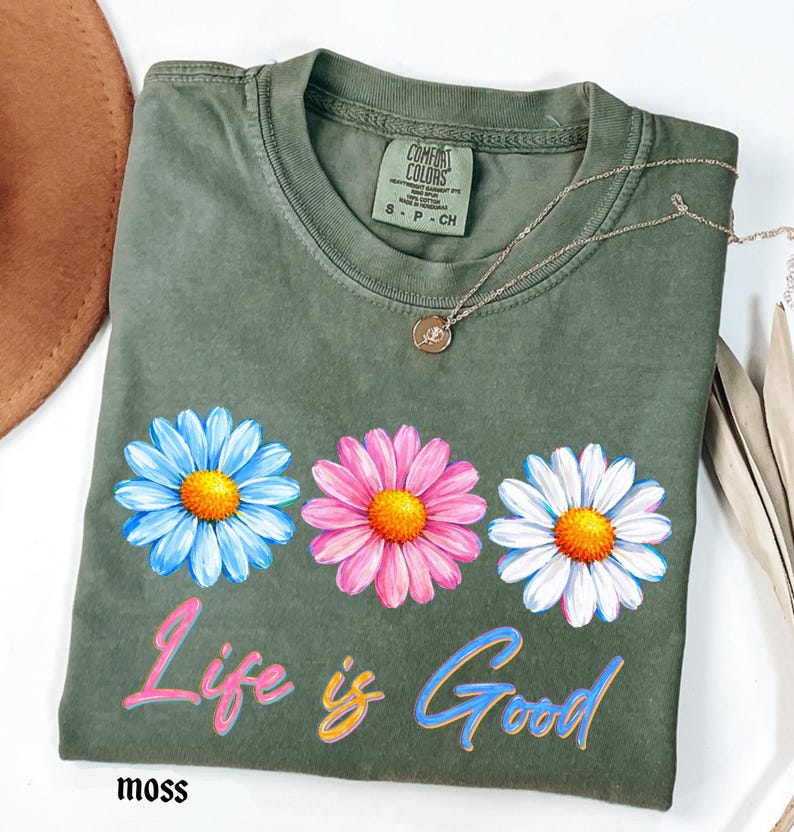 Pode incluir: Camiseta verde musgo com tr&ecirc;s desenhos de flores de margarida em azul, rosa e branco, cada uma com um centro amarelo. A frase "Life is Good" est&aacute; impressa abaixo das flores em uma escrita colorida. Um colar de prata com um pequeno pingente repousa na camisa.