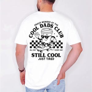Pu&ograve; includere: T-shirt bianco con grafica nera di una tazza di caff&egrave; con occhiali da sole su uno skateboard. Il testo recita "PROUD MEMBER OF THE COOL DADS CLUB" e "STILL COOL JUST TIRED".