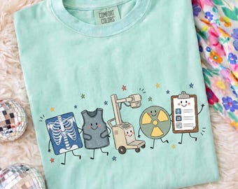 May include: A light mint green t-shirt with a Comfort Colors label at the neck. The graphic features five cartoon characters: an X-ray of a rib cage, a lead apron, an X-ray machine, a radiation symbol, and a clipboard, all with smiling faces.