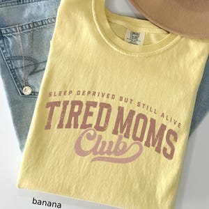 May include: A yellow Comfort Colors t-shirt with the text "SLEEP DEPRIVED BUT STILL ALIVE TIRED MOMS Club" in brown and pink lettering. The shirt is folded on top of blue jeans and a tan hat.