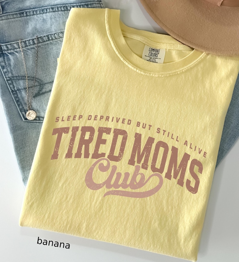 May include: A yellow Comfort Colors t-shirt with the text "SLEEP DEPRIVED BUT STILL ALIVE TIRED MOMS Club" in brown and pink lettering. The shirt is folded on top of blue jeans and a tan hat.