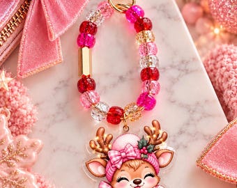 May include: A keychain featuring a gold heart-shaped clasp attached to a loop of alternating pink, red, and clear faceted beads. The charm is a flat acrylic illustration of a reindeer wearing a pink plaid hat and scarf, holding a pink mug.