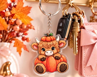 May include: A clear acrylic keyring featuring a cartoon illustration of a fluffy brown Highland cow with a small pumpkin on its head, sitting inside a large orange pumpkin. The keyring has a silver metal split ring and a short chain link.