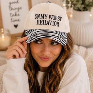 May include: A cream-colored baseball cap featuring the black embroidered text ON MY WORST BEHAVIOR on the front panel. The brim of the cap displays a black and white horizontal striped pattern. The cap has a structured crown and a button on top.