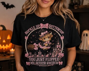 May include: A black short-sleeved t-shirt featuring a graphic of a highland cow wearing a witch hat and riding a broomstick. The design includes pink bows, a castle, tombstones, and the text BUCKLE UP BUTTERCUP YOU JUST FLIPPED MY WITCH SWITCH.