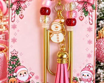 May include: A large gold-tone metal paperclip featuring a dangling charm of a white snowman wearing a pink hat and a pink faux suede tassel. The paperclip is adorned with a beaded loop containing red, pink, and clear faceted beads.