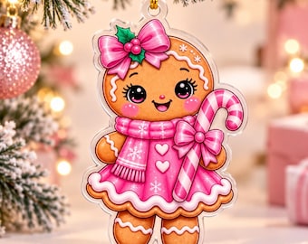 May include: A clear acrylic Christmas ornament shaped like a gingerbread person. The figure wears a bright pink dress with white heart accents, a matching pink scarf with white snowflake patterns, and a large pink bow on its head. It holds a candy cane.