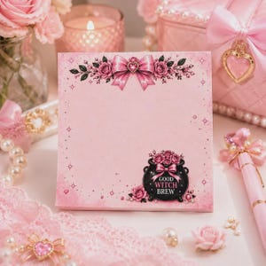 May include: A square pink notepad featuring a decorative pink bow with a heart center at the top and a black cauldron at the bottom right. The cauldron is adorned with pink roses and a pink bow, and it displays the text GOOD WITCH BREW in white.