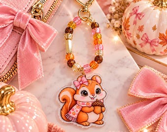 May include: A keyring featuring a heart-shaped gold clasp and a loop of multicoloured beads in shades of pink, orange, and brown. Attached is an acrylic charm of a cartoon squirrel wearing a pink bow and scarf while holding an acorn.
