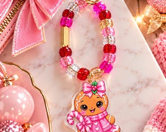 May include: A keyring featuring a heart-shaped gold clasp and a loop of faceted beads in shades of pink, red, and clear. Attached is an acrylic charm of a gingerbread person wearing a pink jumper and bow, holding a candy cane with holly.