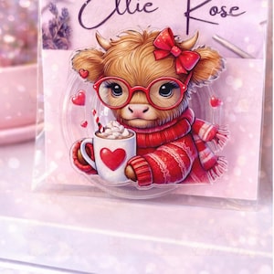 May include: A clear plastic display card featuring the text Ellie Rose and an illustration of a fluffy brown highland cow wearing red glasses, a red bow, and a red sweater, holding a white mug with a red heart and whipped cream.