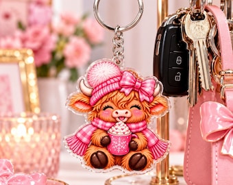 May include: An acrylic keyring featuring a cartoon highland cow wearing a pink knit hat with a pom-pom, a pink plaid scarf, and a matching bow. The cow is holding a pink mug decorated with a white snowflake and heart-shaped foam.