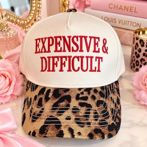 Puede incluir: Una gorra de béisbol de color crema con el texto "EXPENSIVE & DIFFICULT" bordado en rojo en el panel frontal. La gorra tiene una visera curva con un estampado de leopardo en marrón y negro y un pequeño botón oscuro en la parte superior.