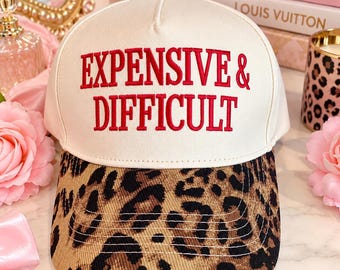 May include: A cream-coloured baseball cap featuring the red embroidered text "EXPENSIVE & DIFFICULT" on the front panel. The cap has a curved brim with a brown and black leopard print pattern and a small dark button at the top centre.