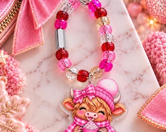 May include: A keyring featuring a silver heart-shaped clasp attached to a loop of faceted beads in shades of pink, red, and clear. An acrylic charm hangs from the beads, depicting a fluffy brown cow wearing a pink beanie and scarf.