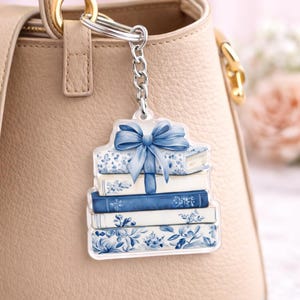 May include: A clear acrylic keychain featuring a stack of books with blue and white floral designs and a blue bow. The keychain is attached to a silver chain and a silver keyring. The background is a beige handbag.