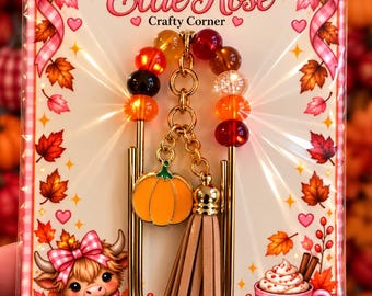 May include: A gold-toned metal jumbo paperclip bookmark featuring a dangling orange enamel pumpkin charm and a beige faux suede tassel. The top of the paperclip is adorned with a row of multicoloured translucent beads in shades of red, orange, and brown.
