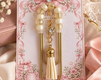 May include: A large gold-tone metal paperclip bookmark adorned with cream-coloured beads, a central gold chain featuring a clear crystal star charm, and a cream-coloured faux leather tassel, all mounted on a decorative pink card.
