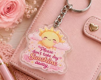 May include: A clear acrylic keyring featuring a smiling yellow sun surrounded by pink clouds and small flowers. The text reads, Please don't take my Sunshine away, in a decorative pink font. The keyring includes a silver metal ring and chain.