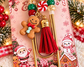 May include: A gold-toned metal paperclip bookmark adorned with a gingerbread man charm, a red suede tassel, and a strand of red, green, and gold glitter beads. A round peppermint candy bead is attached to the top of the metal clip.