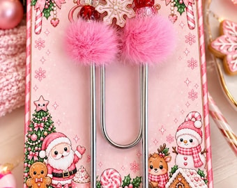May include: A large silver metal paperclip adorned with two pink faux fur pom-poms, red and pink beads, and a white snowflake charm. The paperclip is attached to a rectangular card featuring a festive Christmas illustration of Santa, a snowman, a gingerbread house, and a gingerbread person.
