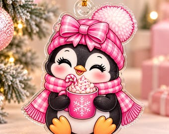 May include: A clear acrylic hanging ornament featuring a black and white penguin wearing a pink plaid knit hat with a pom-pom and a matching pink plaid scarf. The penguin holds a pink mug with a white snowflake design and a candy cane.