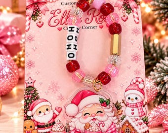 May include: A keychain featuring a gold heart-shaped clasp and a beaded loop with red, pink, and clear beads, including white letter beads spelling HOHO. An acrylic charm depicts a cartoon Santa Claus holding a pink mug with a candy cane.