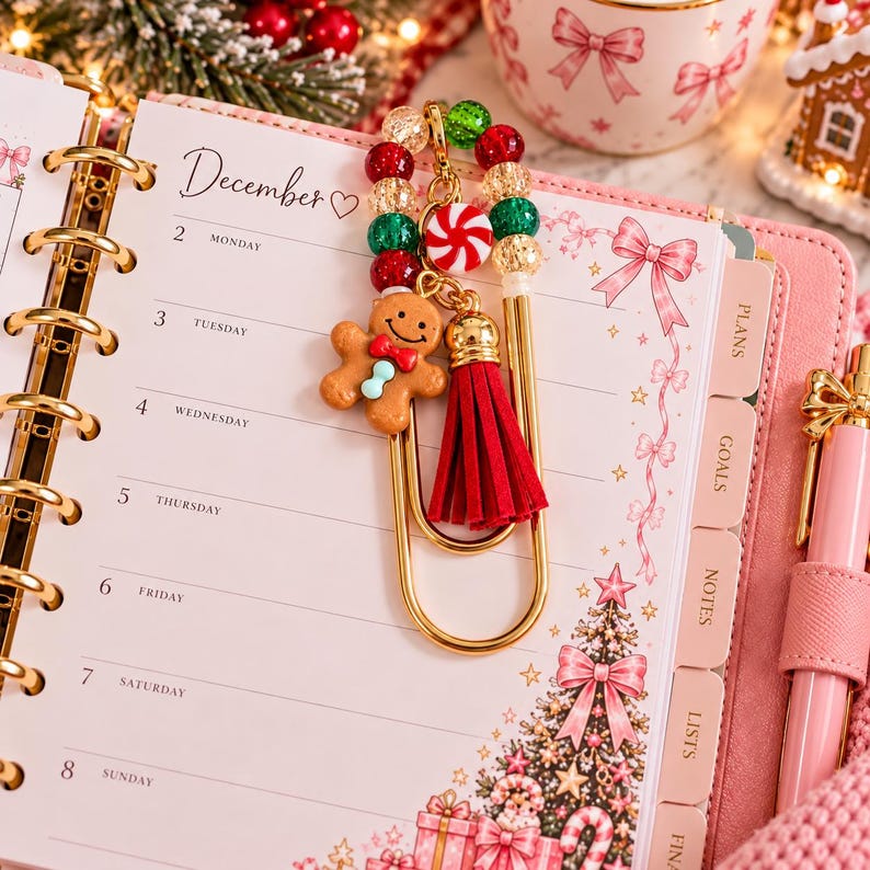 May include: A gold metal paperclip bookmark featuring a gingerbread man charm, a red and white peppermint candy charm, a red suede tassel, and a strand of red, green, and gold glitter beads resting on a December calendar page with holiday graphics.