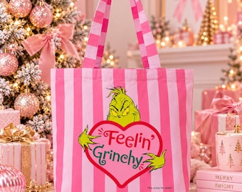 May include: A pink and light pink vertical striped tote bag featuring a graphic of the Grinch character peeking over a red heart. The heart contains the green text Feelin' Grinchy and the Grinch's hands. The bag has two matching striped handles.