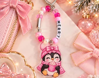 May include: A keyring featuring a heart-shaped silver clasp, a loop of pink, white, and clear beads, and letter beads spelling MERRY. An acrylic charm hangs from the loop, depicting a penguin wearing a pink hat and scarf, holding a pink mug.