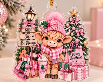 May include: A clear acrylic Christmas ornament featuring a fluffy Highland cow wearing a pink plaid hat and scarf. The design includes a street lamp with signs reading SHOP, SIP, Be Merry, a decorated Christmas tree, gifts, and a pink bag with Merry Christmas text.