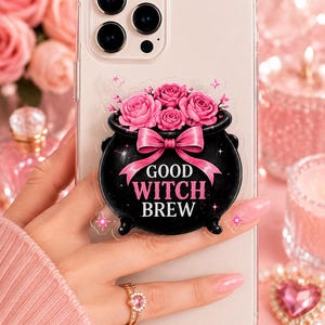May include: A clear smartphone case featuring a black cauldron sticker with pink roses, a pink bow, and the text "GOOD WITCH BREW" in white lettering. The design includes small pink star accents on a transparent background applied to the phone.