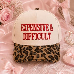 Peut inclure: Une casquette de baseball couleur crème avec le texte EXPENSIVE & DIFFICULT brodé en fil rouge sur le panneau avant. La casquette présente un motif imprimé léopard marron et noir sur la visière et un petit bouton assorti sur le dessus.
