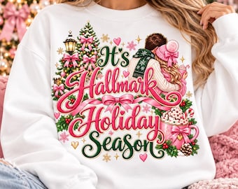 May include: A white crewneck sweatshirt featuring a graphic print of a couple, a decorated Christmas tree, and a mug of hot cocoa. The text reads It's Hallmark Holiday Season in pink and green script, surrounded by pink bows and snowflakes.