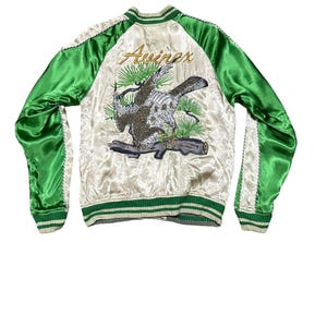 May include: Cream-colored satin bomber jacket with green sleeves and trim. The back features an embroidered design of a bird perched on a branch, with the word "Aviirex" in gold script. The jacket has a ribbed collar, cuffs, and hem.