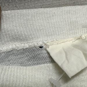 May include: Close-up of white knitted fabric with a textured pattern. A small white tag is attached to the fabric with a plastic fastener. The fabric appears to be part of a garment.