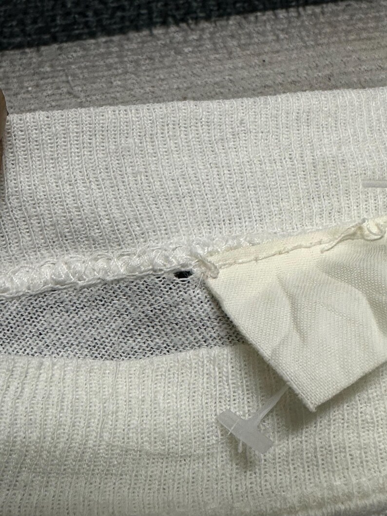 May include: Close-up of white knitted fabric with a textured pattern. A small white tag is attached to the fabric with a plastic fastener. The fabric appears to be part of a garment.