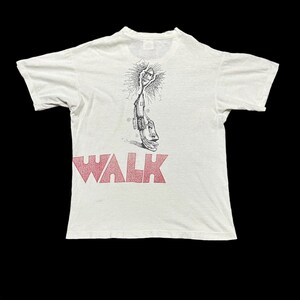May include: White t-shirt with the red word "WALK" at the bottom. The back of the shirt features a black and white illustration of a figure holding a sun-like object. The shirt has short sleeves and a crew neck.