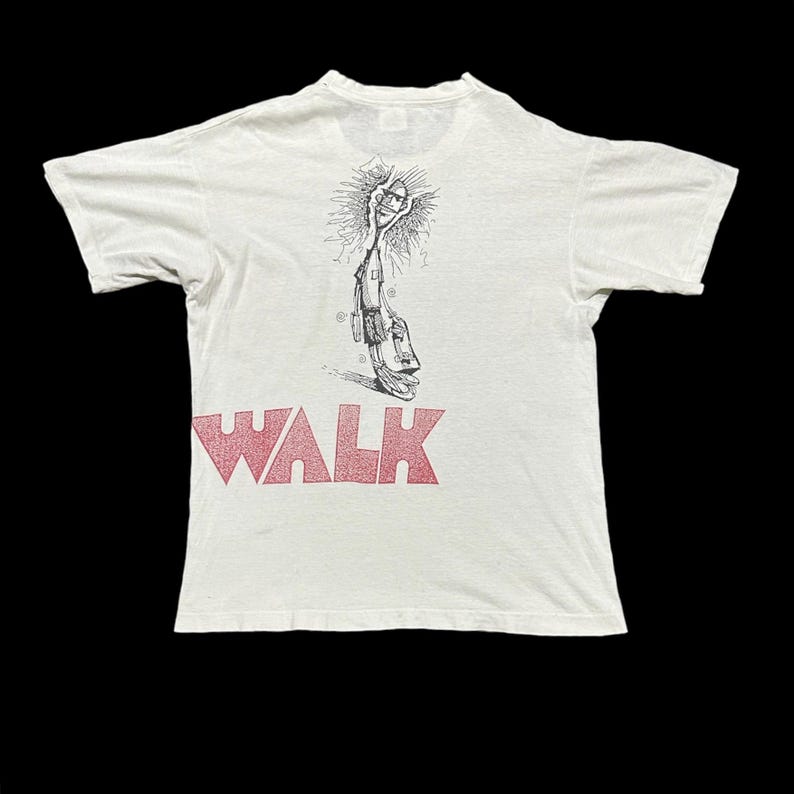 May include: White t-shirt with the red word "WALK" at the bottom. The back of the shirt features a black and white illustration of a figure holding a sun-like object. The shirt has short sleeves and a crew neck.