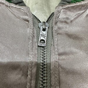 May include: Close-up of a light gray satin jacket with a silver zipper. The zipper pull is marked "JAPAN TOKYO". The collar is trimmed with green and gray stripes. The jacket appears to be vintage.