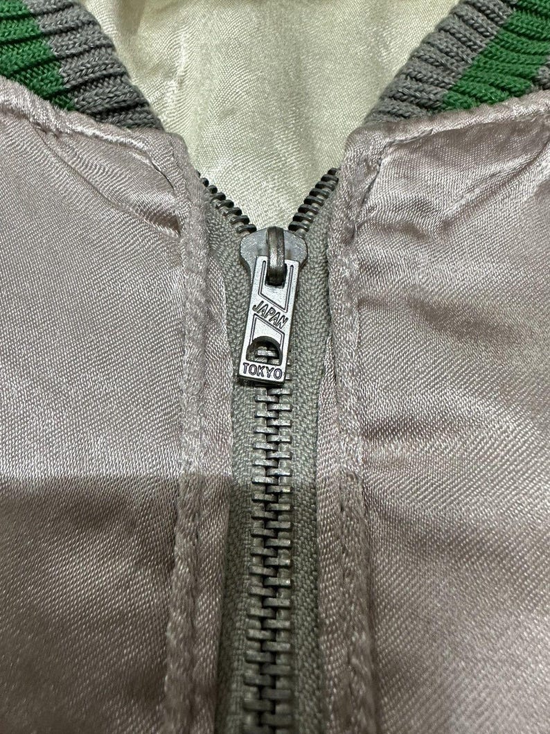 May include: Close-up of a light gray satin jacket with a silver zipper. The zipper pull is marked "JAPAN TOKYO". The collar is trimmed with green and gray stripes. The jacket appears to be vintage.