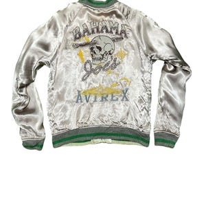 May include: A silver satin bomber jacket with green and gray trim. The back features a skull graphic with the words "Bahama Joes" and "Avirex" in embroidered lettering. The jacket has a ribbed collar, cuffs, and hem.