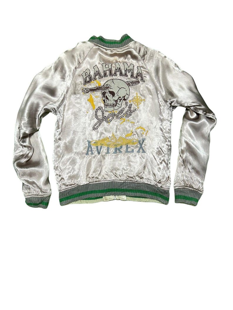 May include: A silver satin bomber jacket with green and gray trim. The back features a skull graphic with the words "Bahama Joes" and "Avirex" in embroidered lettering. The jacket has a ribbed collar, cuffs, and hem.
