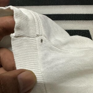 May include: Close-up of a white knitted fabric, possibly a shirt or sweater, showing the texture and seams. The fabric appears soft and has a ribbed detail along the edge.