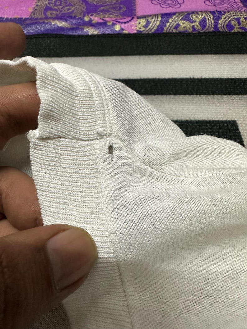 May include: Close-up of a white knitted fabric, possibly a shirt or sweater, showing the texture and seams. The fabric appears soft and has a ribbed detail along the edge.