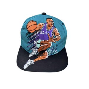 May include: Teal and black baseball cap featuring a basketball player in a purple jersey with the word "SLAM". The cap has a black brim and a black button on top. The basketball player is holding a basketball.